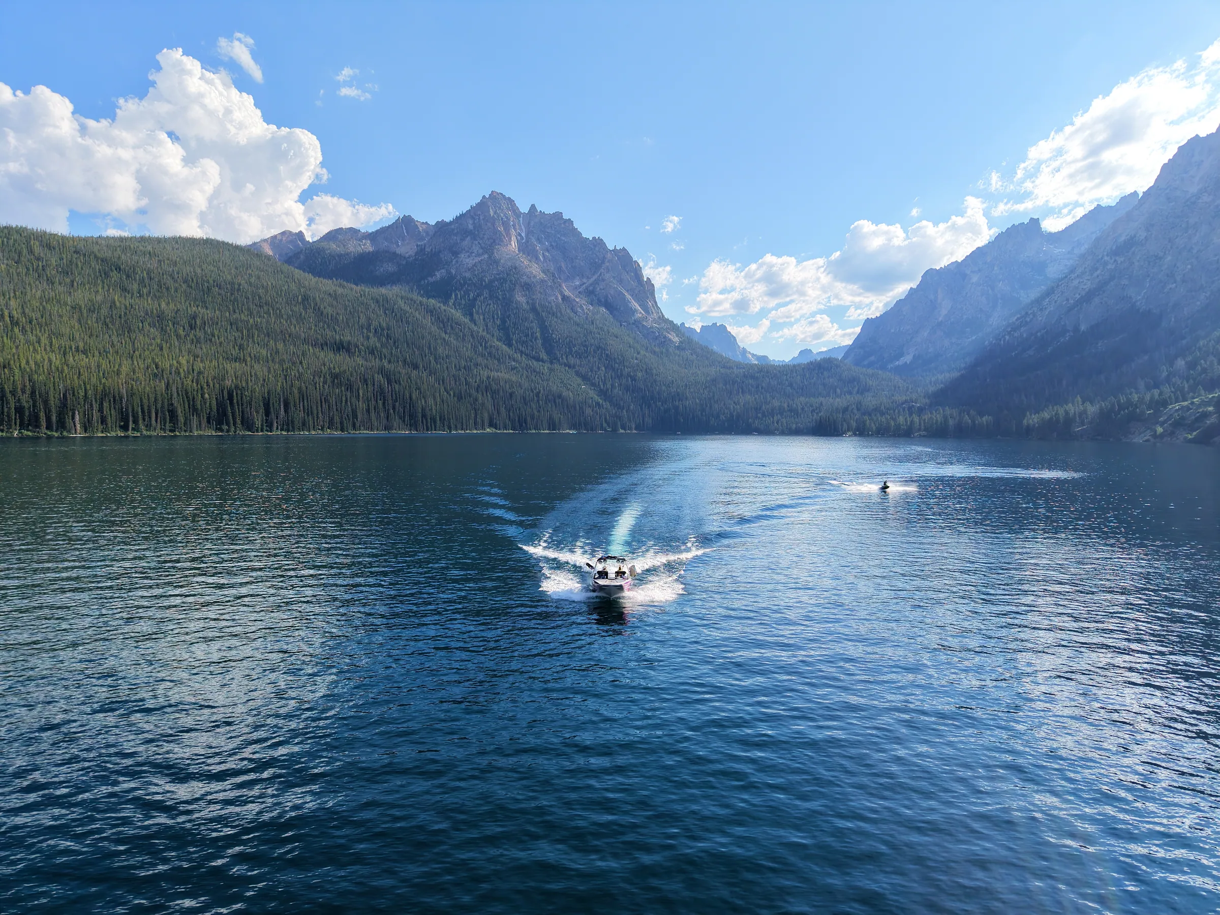 A boat crossing an alpine lake beneath forested mountains and a blue sky.