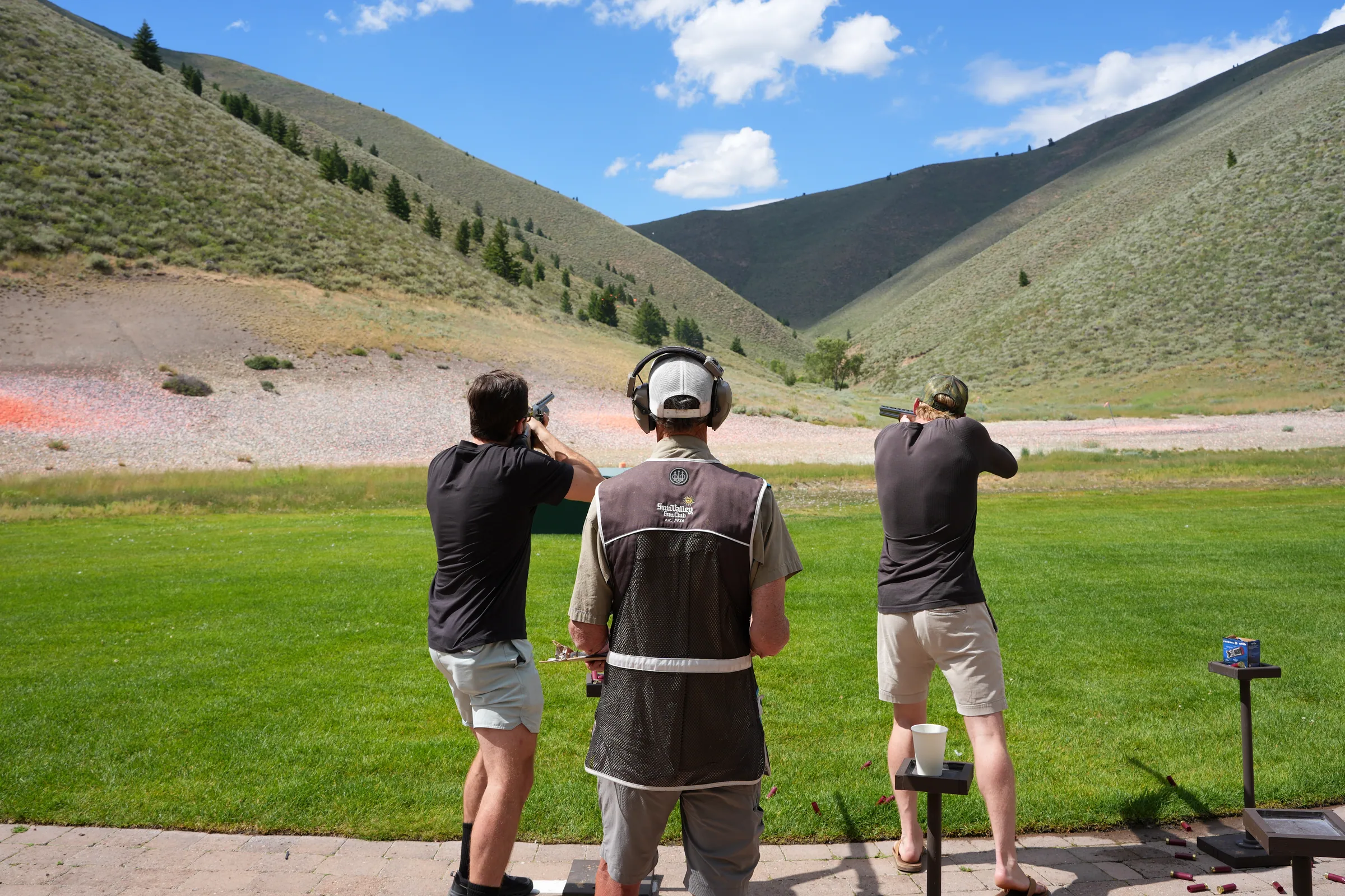 Three adults at an outdoor clay shooting range surrounded by green hills.