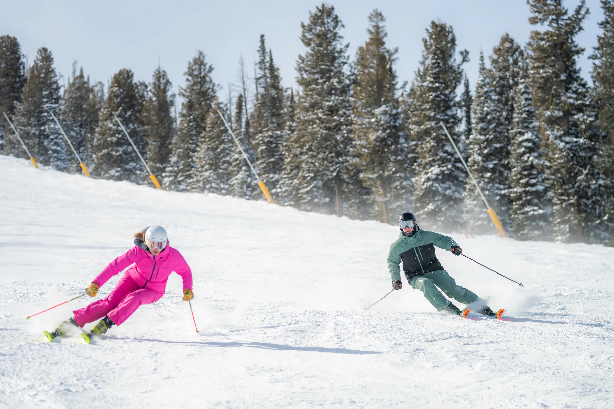 Two skiers carving turns on a snowy slope beside evergreen trees.