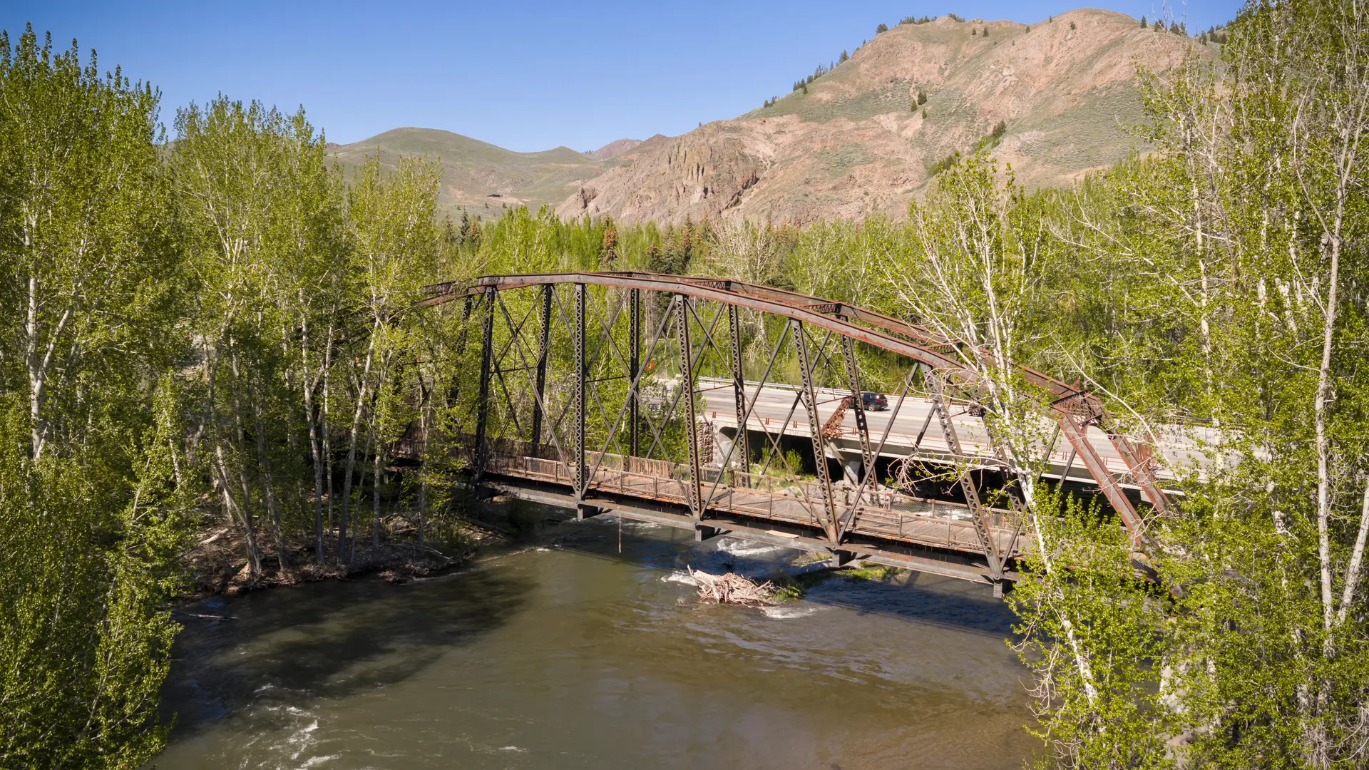 A steel bridge over the river, surrounded by trees and mountain foothills.